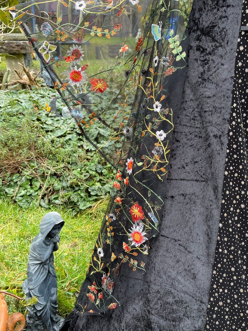 May include: Black sheer fabric with embroidered flowers and a butterfly design. The fabric is draped over a black velvet surface.
