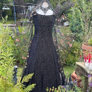 May include: A black velvet gown with a sheer black overlay featuring colorful floral embroidery. The gown has a fitted bodice and a flowing skirt. The gown is displayed on a mannequin in a garden setting.