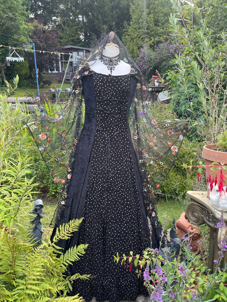 May include: A black velvet gown with a sheer black overlay featuring colorful floral embroidery. The gown has a fitted bodice and a flowing skirt. The gown is displayed on a mannequin in a garden setting.