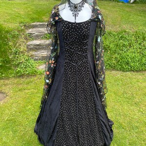 May include: A black velvet gown with a fitted bodice and a full skirt. The gown is adorned with small white sequins and features a lace overlay with floral embroidery. The gown is displayed on a mannequin with a black lace choker.