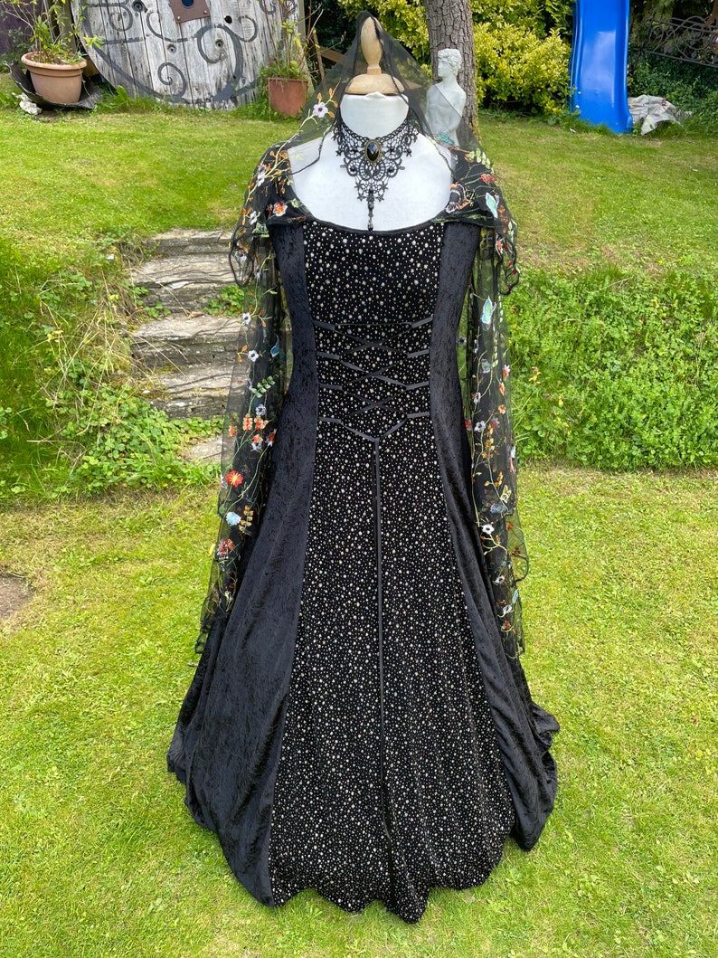 May include: A black velvet gown with a fitted bodice and a full skirt. The gown is adorned with small white sequins and features a lace overlay with floral embroidery. The gown is displayed on a mannequin with a black lace choker.