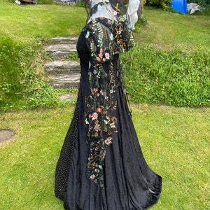 May include: A black velvet gown with a floral embroidered overlay and a sheer black veil. The gown has a fitted bodice and a flowing skirt.