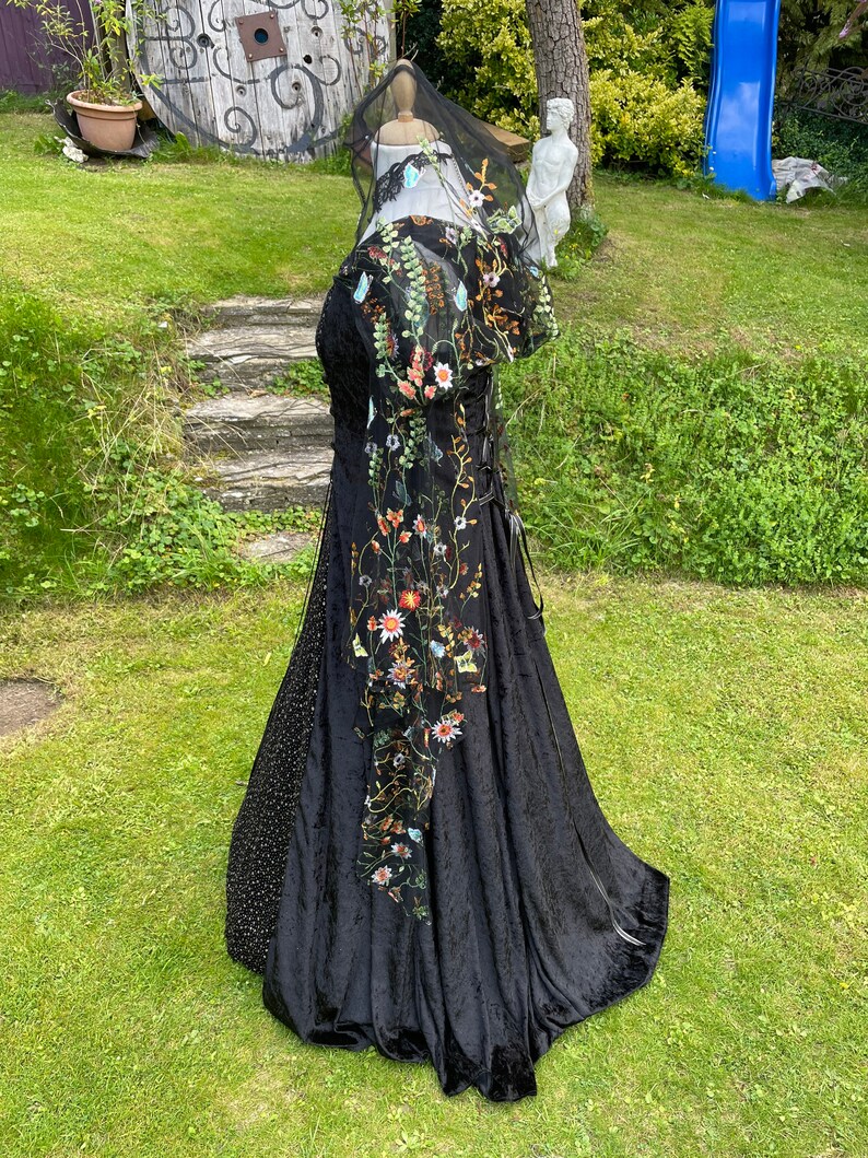 May include: A black velvet gown with a floral embroidered overlay and a sheer black veil. The gown has a fitted bodice and a flowing skirt.
