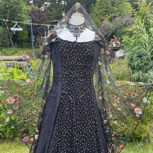 May include: A black velvet gown with a fitted bodice and a full skirt. The gown is adorned with a black lace choker and a sheer black veil with embroidered flowers. The gown is displayed on a mannequin in a garden setting.