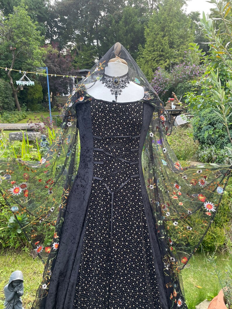 May include: A black velvet gown with a fitted bodice and a full skirt. The gown is adorned with a black lace choker and a sheer black veil with embroidered flowers. The gown is displayed on a mannequin in a garden setting.