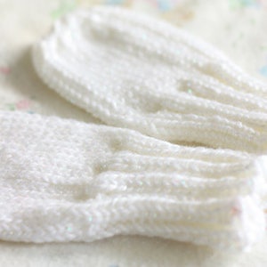 May include: A pair of white knitted baby mittens. The mittens are made of a soft, white yarn and have a simple, classic design.