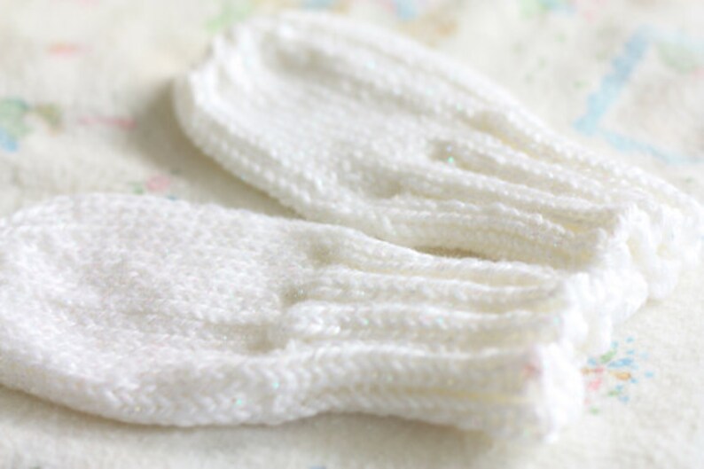 May include: A pair of white knitted baby mittens. The mittens are made of a soft, white yarn and have a simple, classic design.