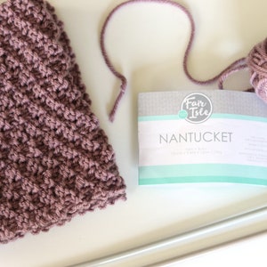 purple knitted seed stitch cowl scarf with knitting needles, ball of yarn, and yarn label