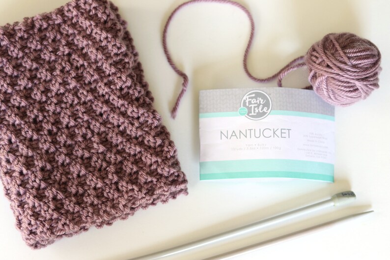 purple knitted seed stitch cowl scarf with knitting needles, ball of yarn, and yarn label