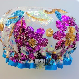 Puede incluir: Un gorro de ducha blanco con un diseño floral hecho de lentejuelas iridiscentes. Las lentejuelas son en tonos de rosa, azul y dorado. El gorro tiene un ribete de satén azul.