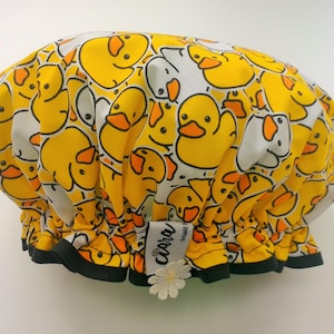 Op de afbeelding: Een gele en witte douchekap met een patroon van cartoon eenden. De douchekap heeft een zwart elastiek en een witte bloem met een groen hart.