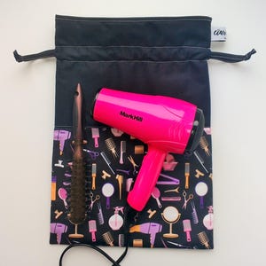 Puede incluir: Una bolsa de cordón negro con un secador de pelo rosa y un cepillo de pelo marrón. La bolsa tiene un patrón blanco y negro de herramientas para peinar el cabello.