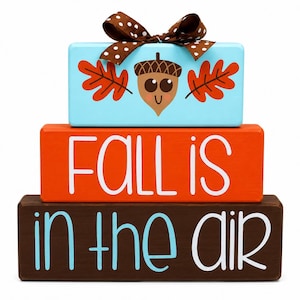 May include: A stack of three rectangular wooden blocks. The top block is light blue with a brown acorn graphic, two orange leaves, and a brown polka-dot ribbon. The middle orange block says FALL IS in white. The bottom brown block says IN THE AIR.