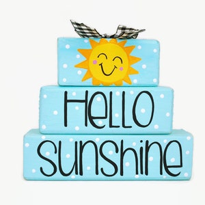 May include: A three-tiered wooden block sign with a blue background and white polka dots. The top block features a yellow smiling sun with a black bow. The middle block says "HELLO" in black lettering. The bottom block says "sunshine" in black lettering.