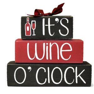 May include: Three wooden blocks stacked on top of each other. The top block is black with white text that says "It's". The middle block is red with white text that says "wine". The bottom block is black with white text that says "o'clock". A red bow is on top of the top block.