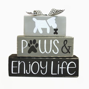 May include: Stacked wooden blocks with a gray and black paint finish. The top block features a white silhouette of a dog with a bone. The middle block says "PAWS &" with a black paw print. The bottom block says "ENJOY LIFE".