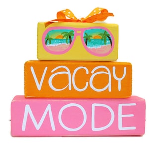 May include: Three wooden blocks stacked on top of each other. The top block is yellow and has a painted image of sunglasses with a beach scene reflected in the lenses. The middle block is orange and has the word "vacay" painted in white. The bottom block is pink and has the word "MODE" painted in white.