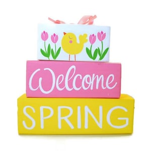 May include: A three-tiered wooden sign with a pink, yellow, and white color scheme. The top tier features a yellow chick with pink tulips and green leaves. The middle tier says "Welcome" in white lettering on a pink background. The bottom tier says "Spring" in white lettering on a yellow background.