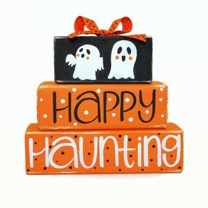 May include: A three-tiered wooden block set with orange and black paint. The top block features two white ghosts with black eyes and mouths. The middle block says "Happy" in white with black polka dots. The bottom block says "Haunting" in white with black polka dots.