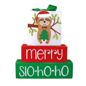 May include: A three-tiered wooden block set with a Christmas sloth design on the top tier. The middle tier reads "merry" in white letters on a red background. The bottom tier reads "slo-ho-ho" in white letters on a green background.