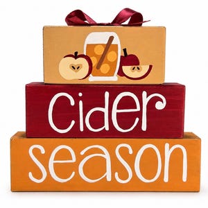 May include: A stack of three rectangular wooden blocks. The top block is tan with a painted glass of cider, apple slices, and a maroon ribbon. The middle block is dark red with the white text "Cider". The bottom block is orange with the white text "season".