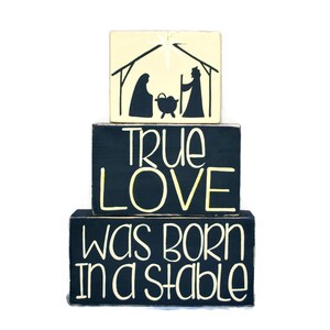 May include: Three wooden blocks stacked on top of each other. The top block is light yellow and features a black silhouette of the nativity scene. The middle block is black with the words "TRUE LOVE" in yellow lettering. The bottom block is black with the words "WAS BORN IN A STABLE" in yellow lettering.