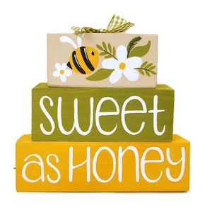 May include: A stack of three rectangular wooden blocks. The bottom block is yellow with the white text "as HONEY". The middle block is olive green with the white text "sweet". The top block is tan with a painted bee, white flowers, and a green gingham bow.