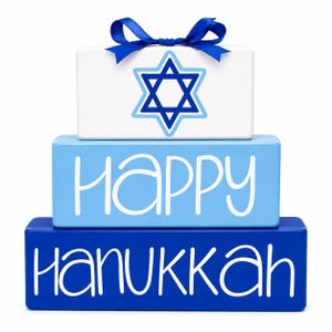 May include: A stack of three rectangular wooden blocks in shades of white, light blue, and dark blue. The top white block features a blue Star of David and a blue ribbon bow. The middle block says HAPPY and the bottom block says HANUKKAH.