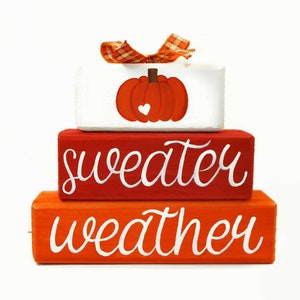 May include: A three-tiered wooden block set with a pumpkin design on the top tier, the middle tier says "sweater" and the bottom tier says "weather". The blocks are painted in shades of orange and red.