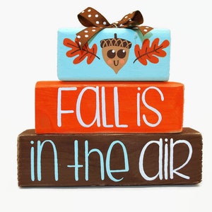 May include: A three-tiered wooden block sign with the words "Fall is in the air" painted in white. The top block features a cartoon acorn with a smiling face and two orange leaves. The blocks are painted in blue, orange, and brown.