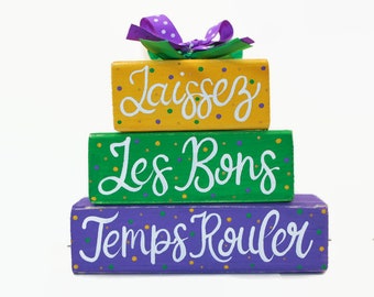 Puede incluir: Un conjunto de bloques de madera de tres niveles con las palabras "Laissez Les Bons Temps Rouler" pintadas en blanco en cada bloque. Los bloques están pintados en morado, verde y amarillo con lunares blancos.