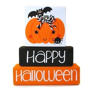 May include: Stacked wooden blocks with a Halloween theme. The top block features a cartoon pumpkin with a bat and spider. The middle block says "Happy" in white letters on a black background. The bottom block says "Halloween" in white letters on an orange background.