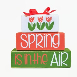 May include: A three-tiered wooden block decoration. The top block is white with red tulip designs and a red bow. The middle block is orange with the word "SPRING" in white. The bottom block is green with the phrase "is in the AIR" in red and white.