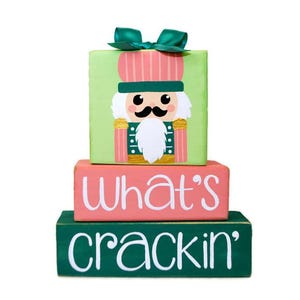May include: A three-tiered wooden block set with a green, pink, and green color scheme. The top block features a painted image of a nutcracker with a pink and green hat. The middle block reads "What's" in white paint. The bottom block reads "crackin'" in white paint.