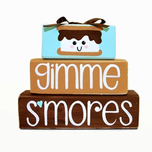 May include: Stacked wooden blocks with a painted image of a s'more on the top block. The middle block says "gimme" and the bottom block says "s'mores".
