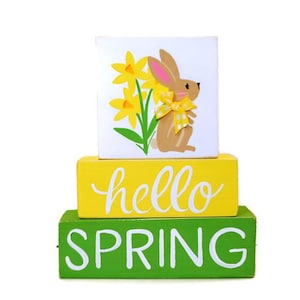 May include: A three-tiered wooden block decoration with a spring theme. The top block features a bunny and daffodils. The middle block says "hello" in white script, and the bottom block says "SPRING" in white block letters. The blocks are painted yellow, green, and white.