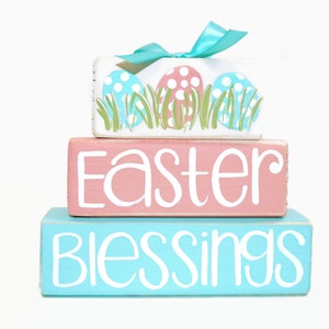 May include: A three-tiered wooden block set with pastel pink, blue, and white paint. The top block features a blue and pink Easter egg design with green grass. The middle block says "Easter" in white paint. The bottom block says "Blessings" in white paint.