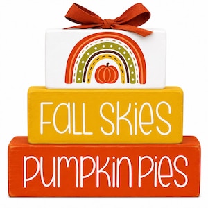 May include: A stack of three rectangular wooden blocks. The top white block features a rainbow and pumpkin graphic with an orange ribbon bow. The middle yellow block displays the white text FALL SKIES. The bottom orange block displays PUMPKIN PIES.