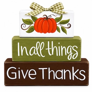 May include: A stack of three rectangular wooden blocks. The top white block features a painted orange pumpkin with green leaves and a green gingham ribbon bow. The middle olive green block says In all things. The bottom brown block says Give Thanks.