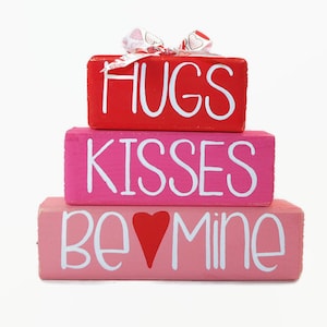 May include: A stack of three wooden blocks with the words "Hugs", "Kisses", and "Be Mine" painted on them in white. The top block is red, the middle block is pink, and the bottom block is pink with a red heart.