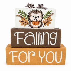 May include: A stack of three rectangular wooden blocks. The top white block features a painted hedgehog holding a pumpkin with autumn leaves and a black and white gingham bow. The middle brown block says Falling and the bottom yellow block says FOR YOU.