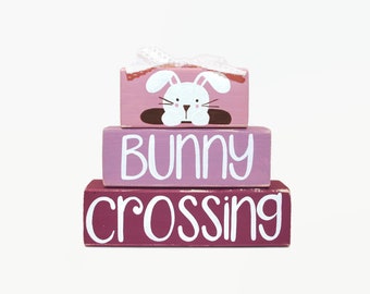 May include: A three-tiered wooden block set with a pink, purple, and burgundy color scheme. The top block features a white bunny with brown ears and a brown nose. The middle block says "Bunny" in white lettering. The bottom block says "crossing" in white lettering.