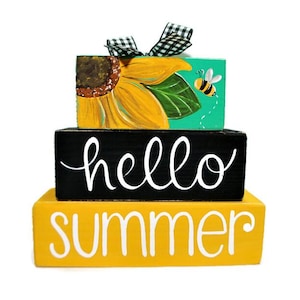 May include: A three-tiered wooden block decoration with a summer theme. The top block features a sunflower and a bee, the middle block says "hello", and the bottom block says "summer". A black and white checkered bow sits on top.