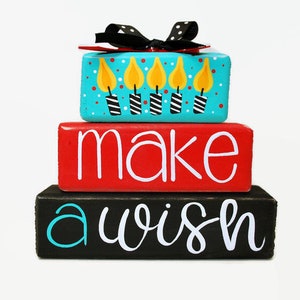 May include: A three-tiered wooden block set with the words "make a wish" painted in white on a black, red, and blue background. The top block features a birthday cake with six candles.