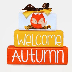 May include: A three-tiered wooden block sign with a cute fox illustration on the top tier, the middle tier says "Welcome" in white lettering on a yellow background, and the bottom tier says "Autumn" in white lettering on an orange background.