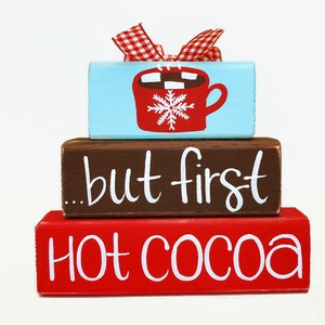 May include: A wooden tiered sign with the words "...but first hot cocoa" painted in white on a brown background. The top tier is painted blue and features a red mug with a snowflake and marshmallows.