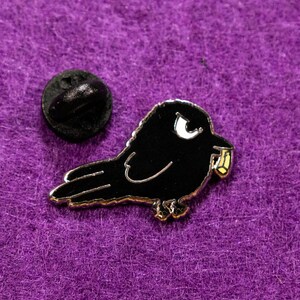 May include: Black enamel pin of a cartoon crow with a white eye and a yellow object in its beak. The pin has a silver finish and a back post for attaching to clothing.