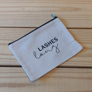 Könnte beinhalten: Eine beige Canvas-Kosmetiktasche mit schwarzem Besatz und dem aufgedruckten Text "LASHES long" in Schwarz.
