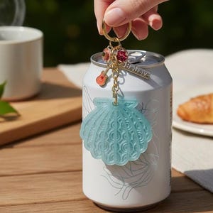 May include: A light blue translucent seashell keychain featuring a gold-tone ring, a small red guitar charm, two round pink crystal beads, and a rectangular silver-tone metal tag engraved with the word Believe, hanging from a white beverage can.