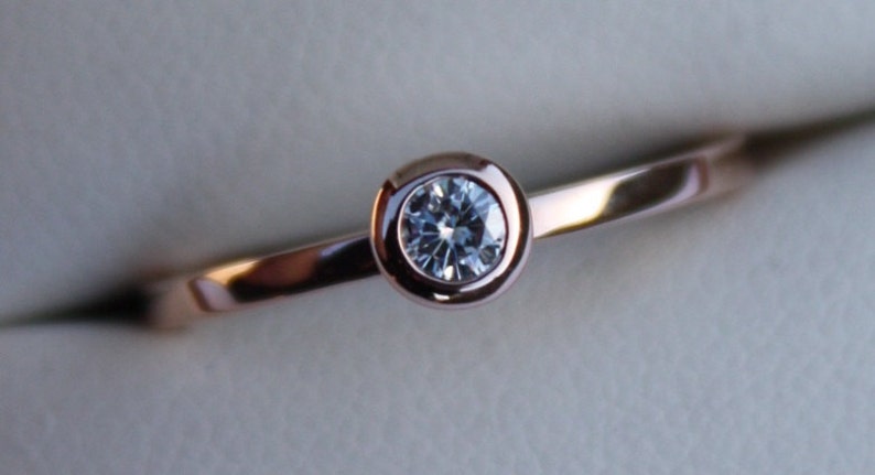 May include: A rose gold ring with a bezel setting featuring a round, clear diamond.
